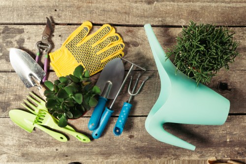 A variety of gardening tools and supplies arranged on a wooden surface, including a small hand trowel, a pair of pruning shears, a pair of yellow gardening gloves, a small green plastic rake, a double-ended cultivator, and a light blue watering can with a round, bushy green plant positioned nearby. The tools appear clean and well-maintained, suitable for general gardening tasks such as planting, pruning, and watering outdoor garden beds or lawn areas typical of residential gardens in Camden Town. The wooden surface provides a rustic background, emphasizing the natural setting. The scene reflects preparation for outdoor garden care, with suitable tools for turf or flower bed maintenance, aligning with professional gardening and landscaping services offered by Landscapers Camden Town in London, in the context of local garden upkeep and lawn care tips.