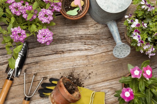 A wooden garden table with various gardening tools and supplies arranged on its surface, including a small trowel, a hand rake, a terracotta pot with soil spilling out, yellow gardening gloves, and a grey watering can. Surrounding the tools are several pots filled with vibrant pink, purple, and white flowering plants, indicating a well-maintained outdoor space typical of Camden Town gardening projects. The weather appears to be bright and clear, with natural light illuminating the scene, emphasizing the textures of the weathered wooden table and the lush greenery and blooms. This setup reflects typical gardening activities such as planting, potting, and outdoor plant care, aligning with the services offered by Landscapers Camden Town, especially within their focus on sustainable gardening practices and recycling. The overall environment suggests a small but diverse garden area, suitable for local garden maintenance and landscape enhancement efforts in the Camden area, postcode NW1.
