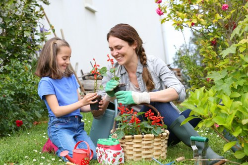 A woman and a young girl are gardening together in a lush back garden in Camden Town, with various green plants, shrubs, and flowering bushes surrounding them. The woman, smiling and wearing gardening gloves, is holding a small potted plant with red flowers, while the girl, also smiling and dressed casually, is assisting by holding the pot. On the grass nearby are gardening tools, a wicker basket, and a decorated watering can, indicating active outdoor garden maintenance or planting. The garden features a combination of neatly trimmed grass, flowering plants, and natural soil beds, creating a vibrant, well-maintained outdoor space suitable for family gardening activities. The environment is bright and sunny, suggesting a clear weather day, highlighting the natural greens, reds, and other earthy tones of the plants and garden furnishings. This scene exemplifies supportive garden care and planting activities often provided by services like those from Landscapers Camden Town, contributing to a beautiful residential outdoor area in the NW1 postcode district.