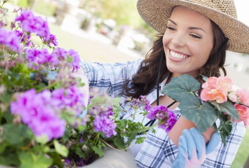 A woman with long brown hair wearing a wide-brimmed straw hat, a blue and white checkered shirt, and light blue gardening gloves is smiling while tending to colorful flowering plants in a well-maintained garden. The garden features a lush green lawn in the foreground, bordered by a variety of vibrant pink, purple, and peach flowers, with some green foliage and shrubs visible. In the background, there are trees and a sunny sky, indicating a bright, pleasant day. The scene reflects a tidy outdoor space where garden maintenance and planting activities are taking place, typical of professional gardening services in Camden Town, London, highlighting the care of outdoor landscaped areas.
