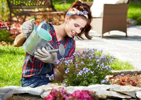 A young woman with brown hair tied back and wearing a colorful plaid shirt, denim jeans, and gardening gloves, is kneeling on the grass in a well-maintained garden in Camden Town. She is holding a metal watering can in one hand and a trowel in the other, actively tending to a small flower bed filled with purple and pink flowering plants. The garden features a lush green lawn in the foreground, bordered by a low stone edging, with neatly arranged flower beds and a variety of shrubs and small trees in the background. The setting appears to be an outdoor space on a sunny day, with natural light illuminating the vibrant colours of the flowers and foliage, and a patio area with patio furniture visible in the background. The scene showcases elements of gardening and outdoor maintenance, aligning with services offered by Landscapers Camden Town to enhance and care for residential landscaped gardens and outdoor spaces in the local area.