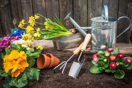 A landscaped garden area featuring a freshly tilled flower bed with rich, dark soil, bordered by a weathered wooden fence in the background. The garden includes vibrant flowering plants such as primroses in bright pink, yellow, and purple shades, along with lush green foliage, creating a colourful display. In the foreground, gardening tools including a hand trowel and a small rake are resting on the soil, alongside a classic metal watering can with a point-of-use spout. A terracotta plant pot lies on its side, indicating recent planting activity. To the side, there are additional flower pots, some filled with blooming flowers, ready for planting or re-potting. The overall scene suggests active garden maintenance suitable for gardening services in Camden Town, with a focus on flower bed preparation and plant care. The setting appears to be outdoors on a bright day, highlighting natural light that enhances the vivid colours of the flowers and the textures of the tools and soil, supporting professional gardening and landscaping efforts in the area.
