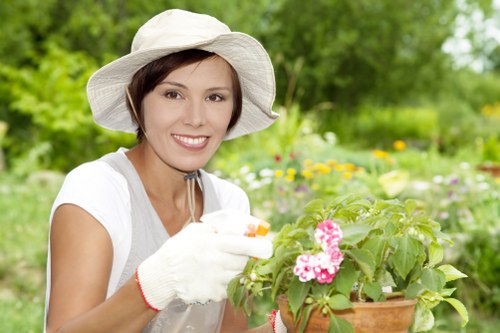 A woman in a white gardening hat and gloves is smiling while tending to a potted plant with pink flowers in a garden setting. Behind her, there is a well-maintained outdoor space featuring a lush green lawn, various shrubbery, and trees that create a natural backdrop. The garden appears to be in full bloom with colourful flowers and dense foliage, indicating healthy, well-cared-for plants. The lighting suggests a bright, mild day, enhancing the vibrant natural tones of the garden. In the foreground, the gardener holds the pot with both hands, showing careful attention to her outdoor planting activities. The scene reflects a peaceful, landscaped yard that may include a variety of plant types, with neatly arranged garden beds and a mix of textures from the flowering plants and the soft grass area. This image aligns with gardening and outdoor maintenance services offered by Landscapers Camden Town, contributing to a visually appealing and thriving garden environment in Camden Town or nearby areas.