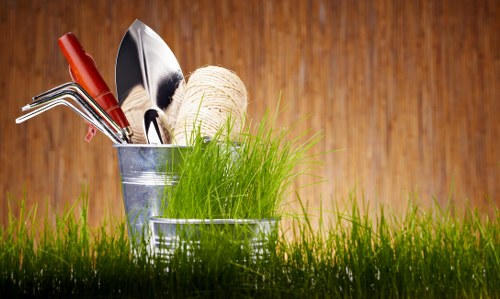 A close-up image of a gardening scene featuring a small metal bucket filled with gardening tools, including a trowel, a cultivator, and a pair of pruning shears, alongside a ball of twine and a clump of fresh, green grass. The bucket is placed on a lush, vibrant grassy lawn with dense, healthy grass blades in the foreground. The background reveals a warm-toned wooden garden fence, softly illuminated by natural daylight, suggesting a tidy and well-maintained outdoor space suitable for planting and landscaping activities in Camden Town. The arrangement reflects gardening preparations and outdoor maintenance work typical of professional landscaping services offered by Landscapers Camden Town, emphasizing readiness for planting or lawn care tasks in a residential garden environment.
