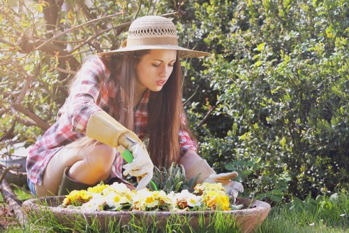 A young woman with long brown hair wearing a straw sunhat, red plaid shirt, and gardening gloves is kneeling in a lush, well-maintained garden in Camden Town, tending to a flower bed filled with yellow, white, and purple blooms. The garden features a neatly edged lawn with dense hedge borders and a variety of green shrubs and trees in the background, creating a vibrant and natural outdoor environment. Sunlight filters through the foliage, casting a warm glow over the scene, highlighting the textures of the grass, soil, and plants. This setting reflects the type of outdoor space that landscaping and gardening services by Landscapers Camden Town might help maintain, including flower bed planting, hedge trimming, and lawn care, ensuring a visually appealing and healthy garden in a residential area close to Camden Town.