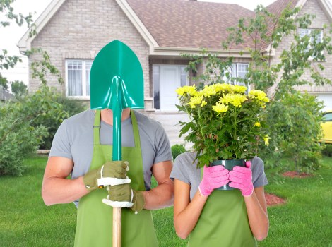 A gardener and a woman are standing on a lush, well-maintained lawn in a residential garden in Camden Town, with a semi-detached brick house in the background. The gardener, wearing a light grey t-shirt, a green apron, and protective gloves, is holding a blue-handled shovel with its blade buried in the ground. The woman, dressed in a grey t-shirt and pink gardening gloves, is covering her face with a large potted plant filled with bright yellow flowers, possibly chrysanthemums or daisies, in a pink container. The garden features neatly trimmed shrubs, a small tree with emerging green leaves, and a variety of plants along the perimeter. The grass is dense and evenly cut, and the overall scene suggests outdoor maintenance and garden enhancement activities suitable for a professional gardening service operating in the Camden Town area, emphasizing the natural tones and landscaping details of a typical suburban garden under daylight conditions.