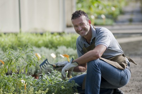 A man in casual clothing, including a grey T-shirt and jeans, is kneeling on the ground in a garden, smiling at the camera while holding a small hand rake. The garden features a lush, green lawn in the foreground with a variety of plants and flowering shrubs alongside the border. Behind him, there are wooden fencing panels and a row of trees or tall bushes providing background privacy, suggesting a residential outdoor space in Camden Town. The soil appears freshly tilled or well-maintained, with some patches of dirt visible among the plants. Natural sunlight illuminates the scene, indicating a clear weather day, and highlighting the vibrant green hues and natural textures of the foliage. This scene reflects typical garden maintenance activities, and the presence of gardening tools and plants emphasizes ongoing landscaping or gardening work, resonating with professional outdoor garden services offered by companies such as Landscapers Camden Town, especially within the context of a garden renovation project in the local area.
